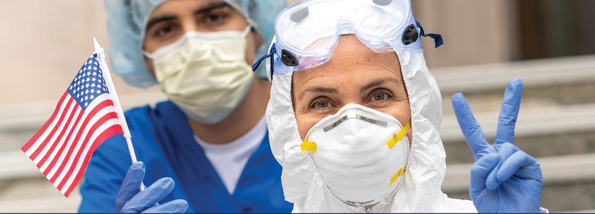 Two health workers in PPE - one holds mini American flag, the other holds up peace or victory sign