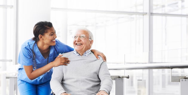 A smiling, female health care worker wearing scrubs stands next to a smiling, elderly male patient, with her arm around his shoulder.