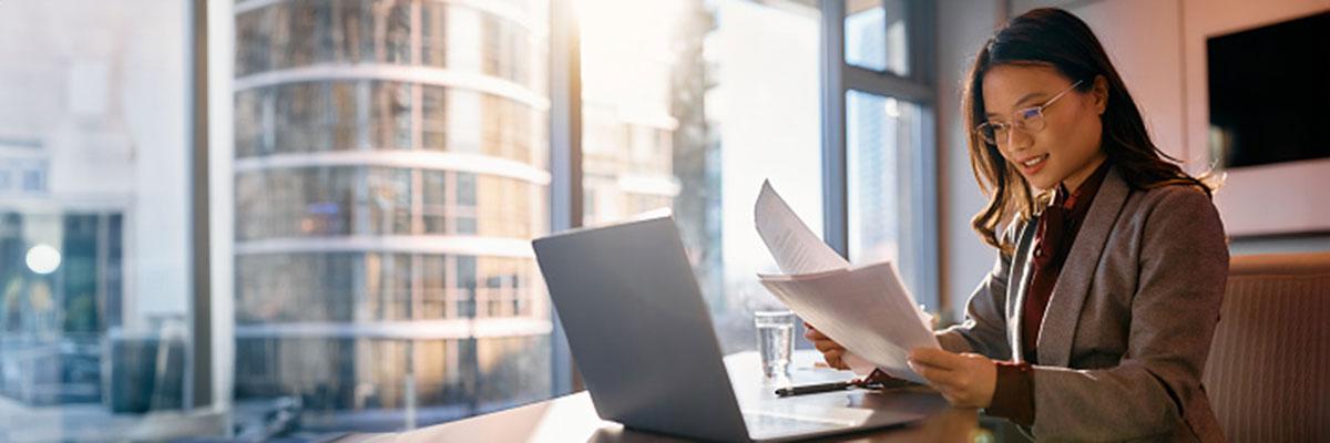 Woman sits in office at desk reading papers, another office building visible through a window in the background